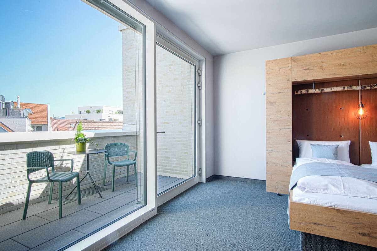 A hotel room with a large window, a balcony with chairs, a bed with a wooden headboard, and a potted plant.