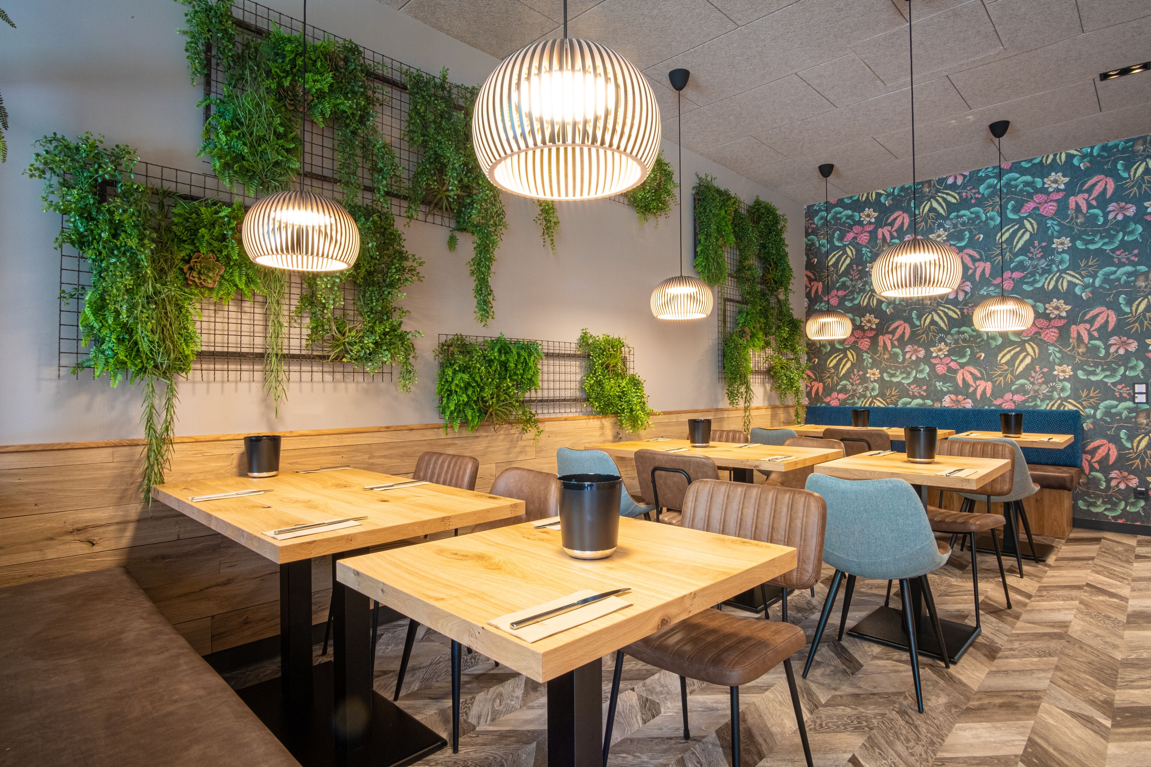 Un interior de restaurante moderno con mesas de madera, sillas de metal negro, lámparas colgantes y plantas verdes en las paredes.