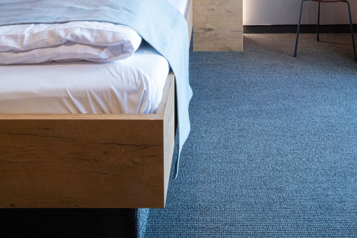 A modern hotel room with a wooden headboard, a large bed with white linens, a round wall-mounted light fixture, and a small stool.