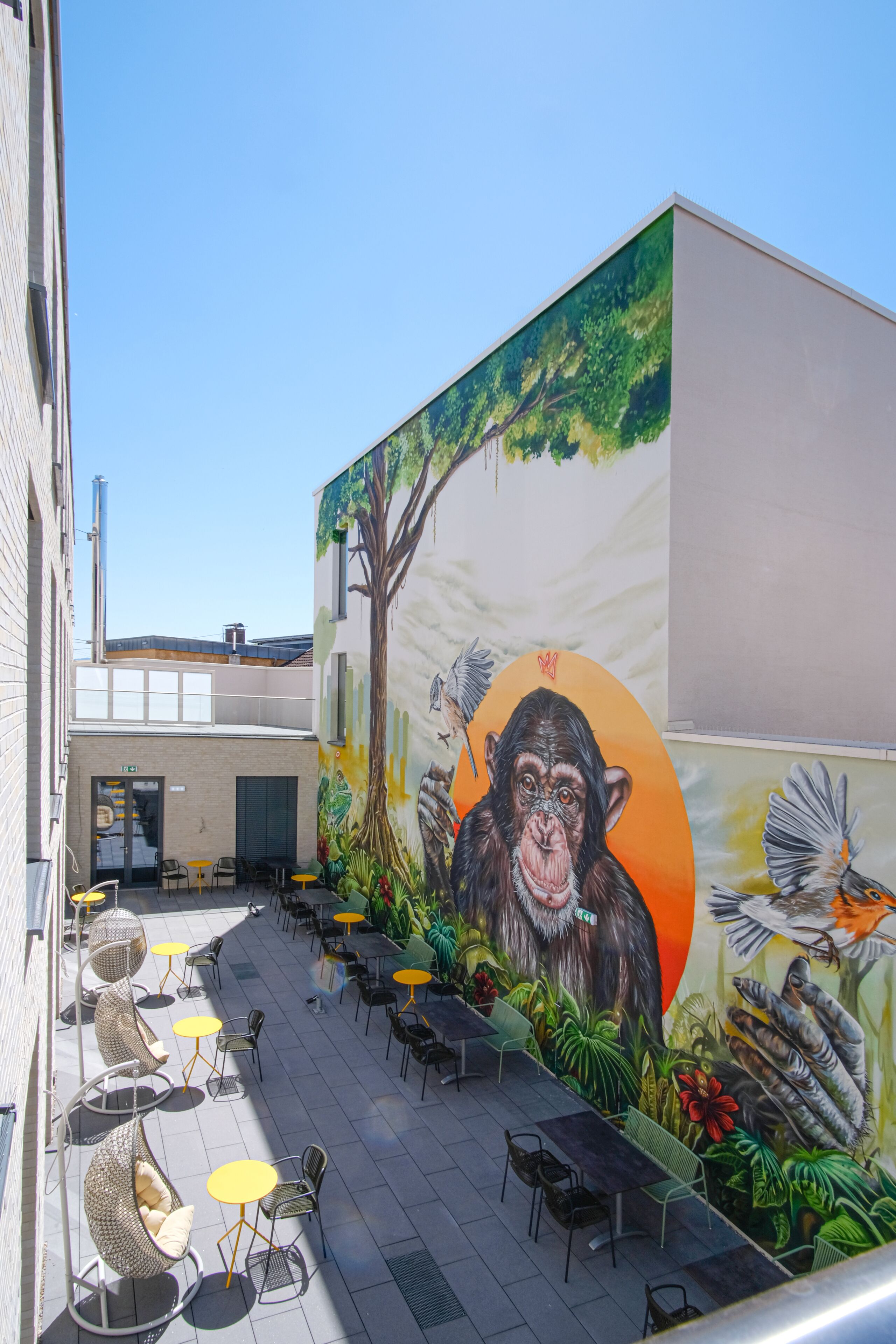 An outdoor seating area with tables and chairs, a mural of a monkey and birds, and a building with a door and windows.
