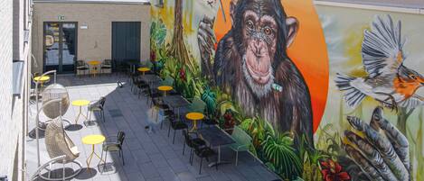 An outdoor seating area with tables and chairs, a mural of a monkey and birds, and a building with a door and windows.
