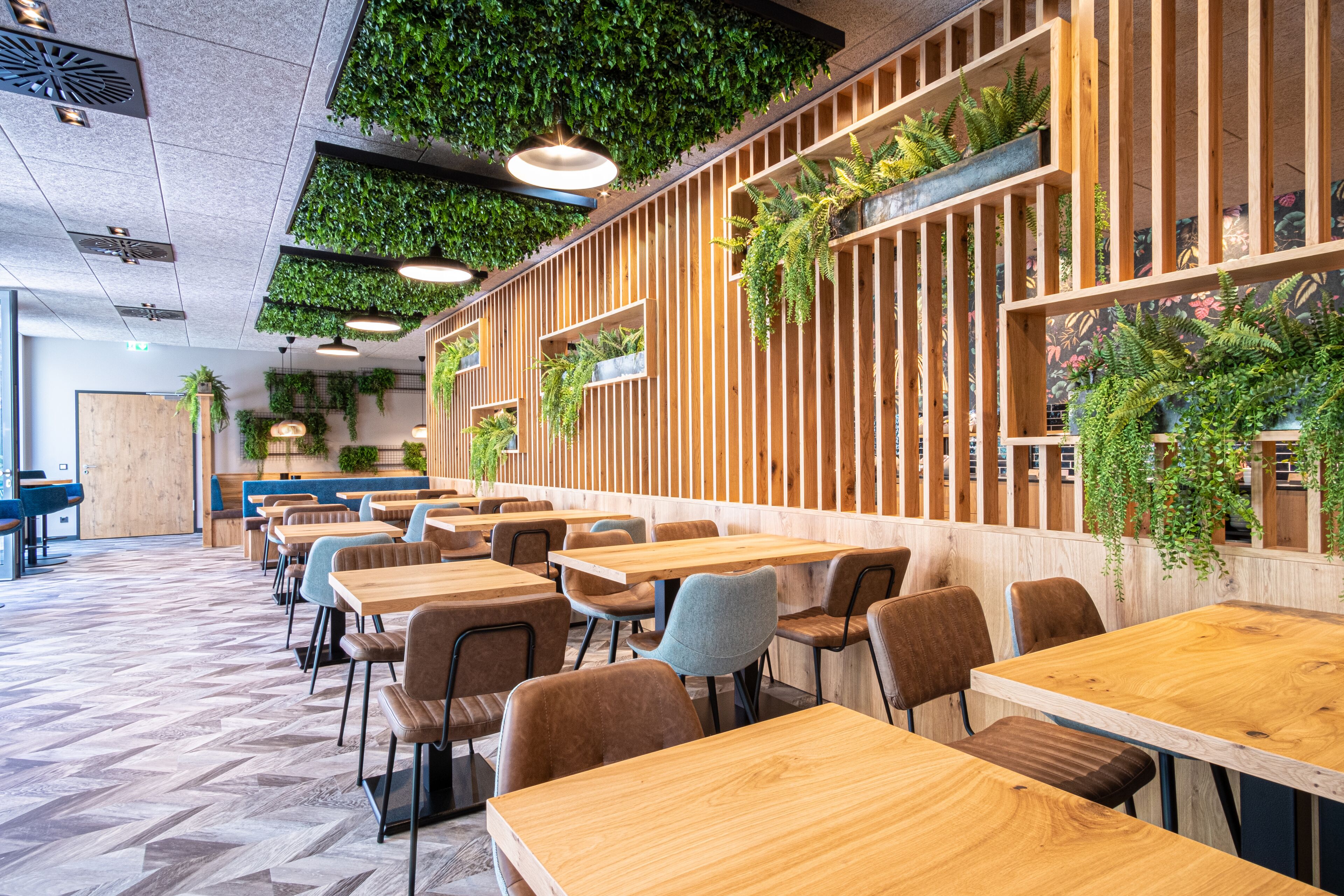 Un interior de restaurante moderno con mesas y sillas de madera, plantas verdes en las paredes y un patrón de suelo en espiga.
