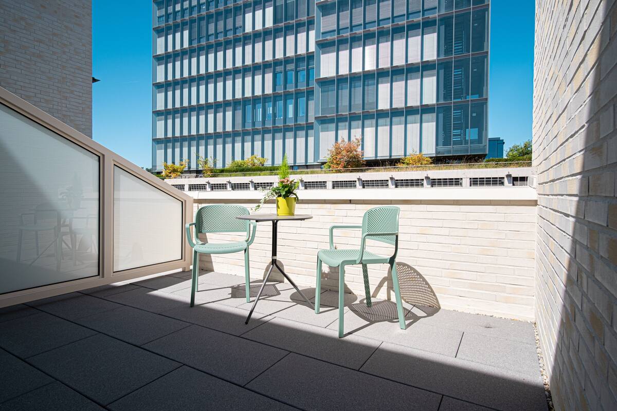 A balcony with two green chairs and a small table, overlooking a modern building with large windows.