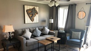 Una sala de estar con un sofá gris, una mesa de centro, una mesita de noche con una lámpara, un sillón azul y un ventilador de techo.