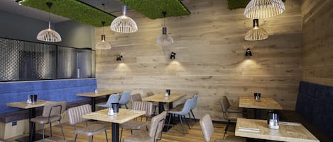A modern restaurant interior with wooden flooring, green artificial grass ceiling panels, and pendant lighting. Tables and chairs are arranged for dining.