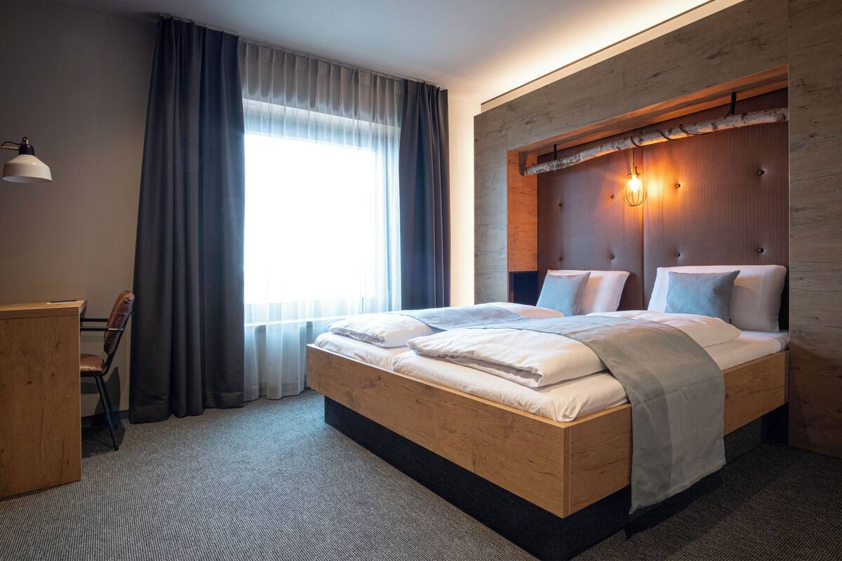 A modern hotel room with a wooden bed, a desk, and a large window with curtains.
