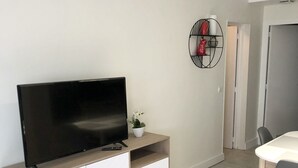 Ein modernes Wohnzimmer mit Fernseher auf einer Holzkommode, einer kleinen Topfpflanze und einem Essbereich mit Tisch und Stühlen.