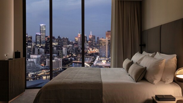 A modern hotel room with a large bed, a nightstand, and a view of a cityscape at dusk. - Oakwood Premier Melbourne (Melbourne)