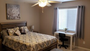 Ein Schlafzimmer mit Bett, Nachttischen, einem Schreibtisch mit Stuhl, einem Deckenventilator, einem Fenster mit Vorhängen und einem Wandbild.
