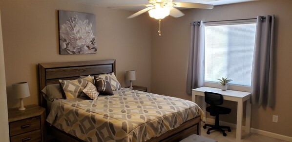 A bedroom with a bed, bedside tables, a desk with a chair, a ceiling fan, a window with curtains, and a wall-mounted painting.