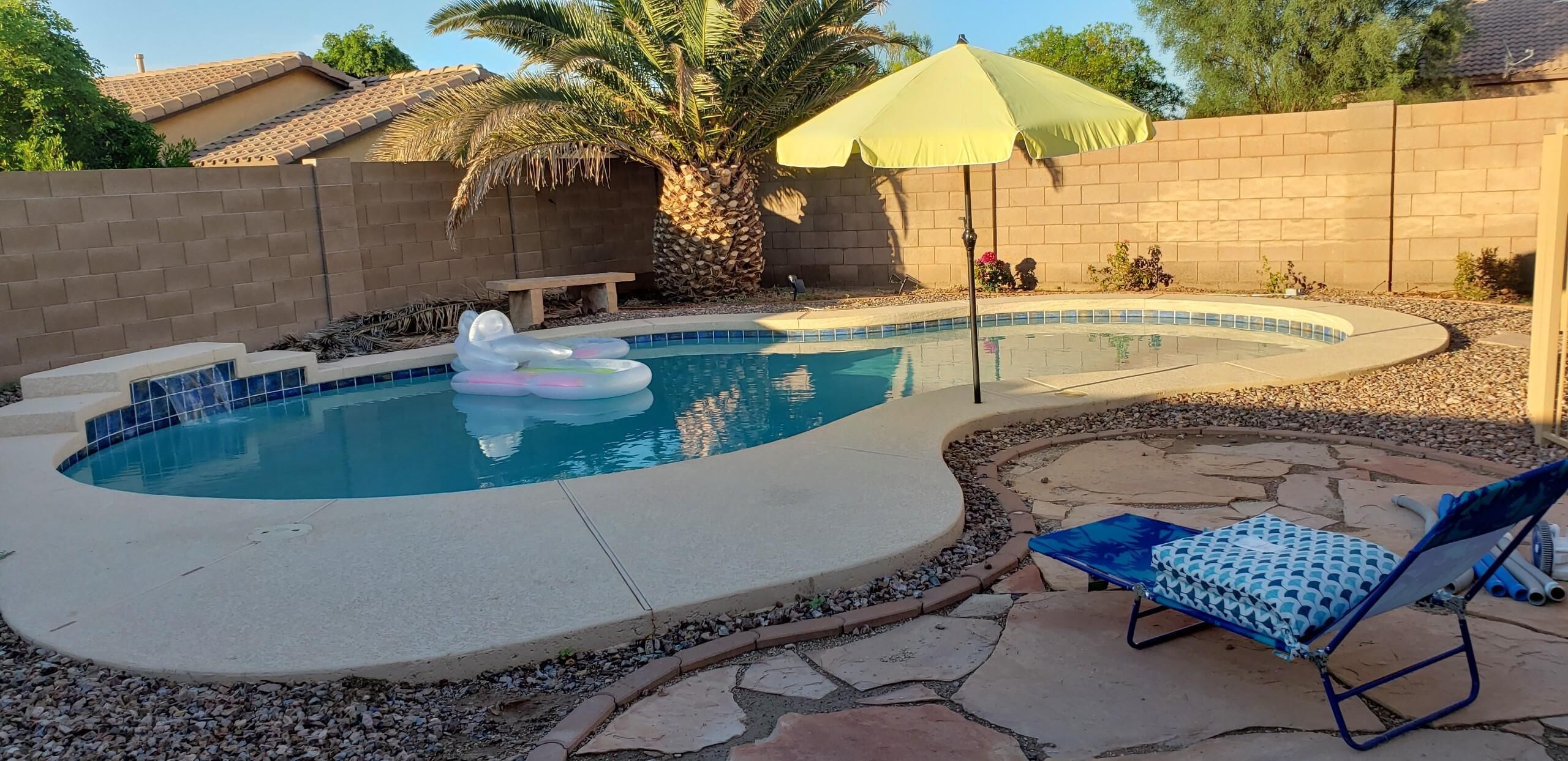 A backyard with a swimming pool, inflatable floaties, a lounge chair, and a palm tree.