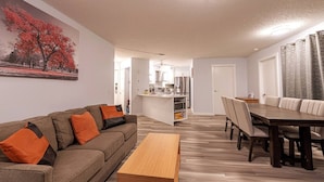 Ein Wohnzimmer mit einer Couch, einem Holztisch, einem Esstisch mit Stühlen, einer Küchenzeile und einem Flur, der in ein weiteres Zimmer führt.