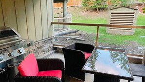 En udendørs terrasse med grill, fletmøbler og et glasbord.