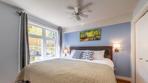 Ein Schlafzimmer mit Bett, Deckenventilator, Fenster mit Vorhängen, einem Bild an der Wand und einer Nachttischlampe.