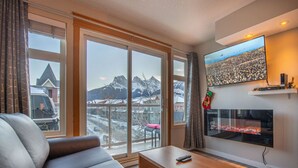 A living room with a sofa, a coffee table, a fireplace, and a large window with a mountain view. - Top-Floor Condo | 2BR + AC with Mountain Views (Canmore)
