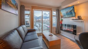 A modern living room with a leather sofa, a wooden coffee table, a fireplace, and a large window overlooking a snowy mountain landscape. - Top-Floor Condo | 2BR + AC with Mountain Views (Canmore)