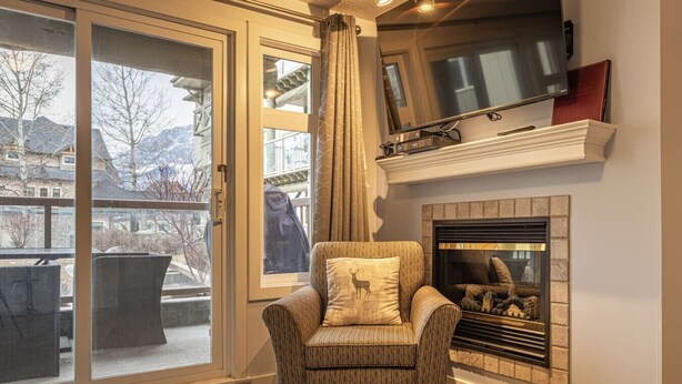 A living room with a fireplace, a television mounted above it, and a large window with a view of buildings and trees. - Mountain View/Full Kitchen/BBQ/AC/King Bed (Canmore)