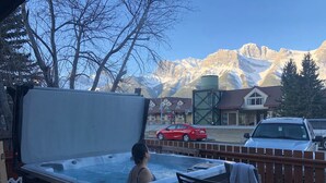 Ein Whirlpool mit einer Person darin, eine Holzterrasse, Blick auf Berge und ein rotes Auto in der Nähe.