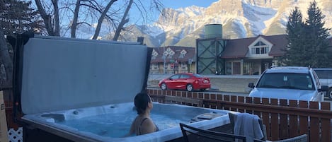 Ett bubbelbad med en person i, en träterrass, utsikt över berg och en röd bil parkerad i närheten.