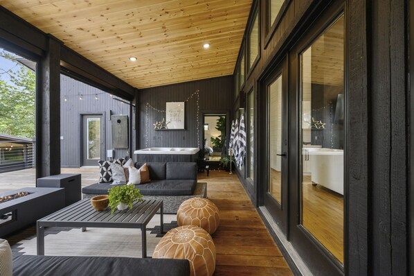 Un espace de vie extérieur moderne, doté d’un plafond en bois, d’un ensemble de canapés, d’un foyer et offrant une vue sur la cuisine grâce à de grandes fenêtres.