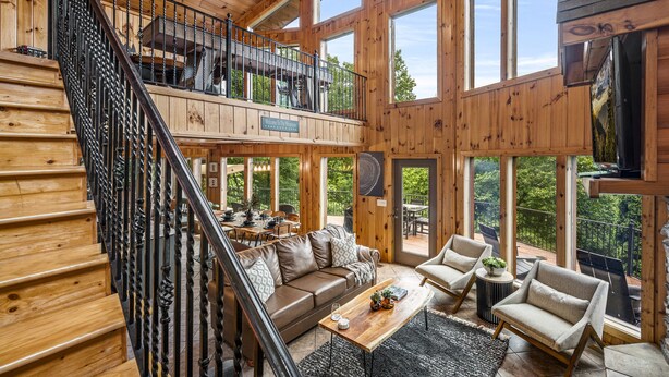 Un intérieur spacieux en bois, doté d’un escalier, d’un coin salon avec un canapé et des fauteuils, et de grandes fenêtres offrant une vue sur l’extérieur.