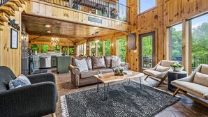 Une cabane en bois spacieuse, avec un espace de vie comprenant un canapé, des fauteuils et une table basse. On aperçoit une cuisine au fond, et il y a un balcon avec une balustrade.