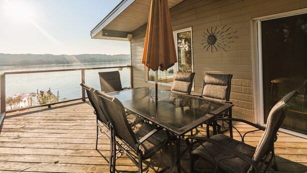 A deck with a glass table and chairs, overlooking a body of water and hills. - Chateau Champlain Cottage (Penetanguishene)