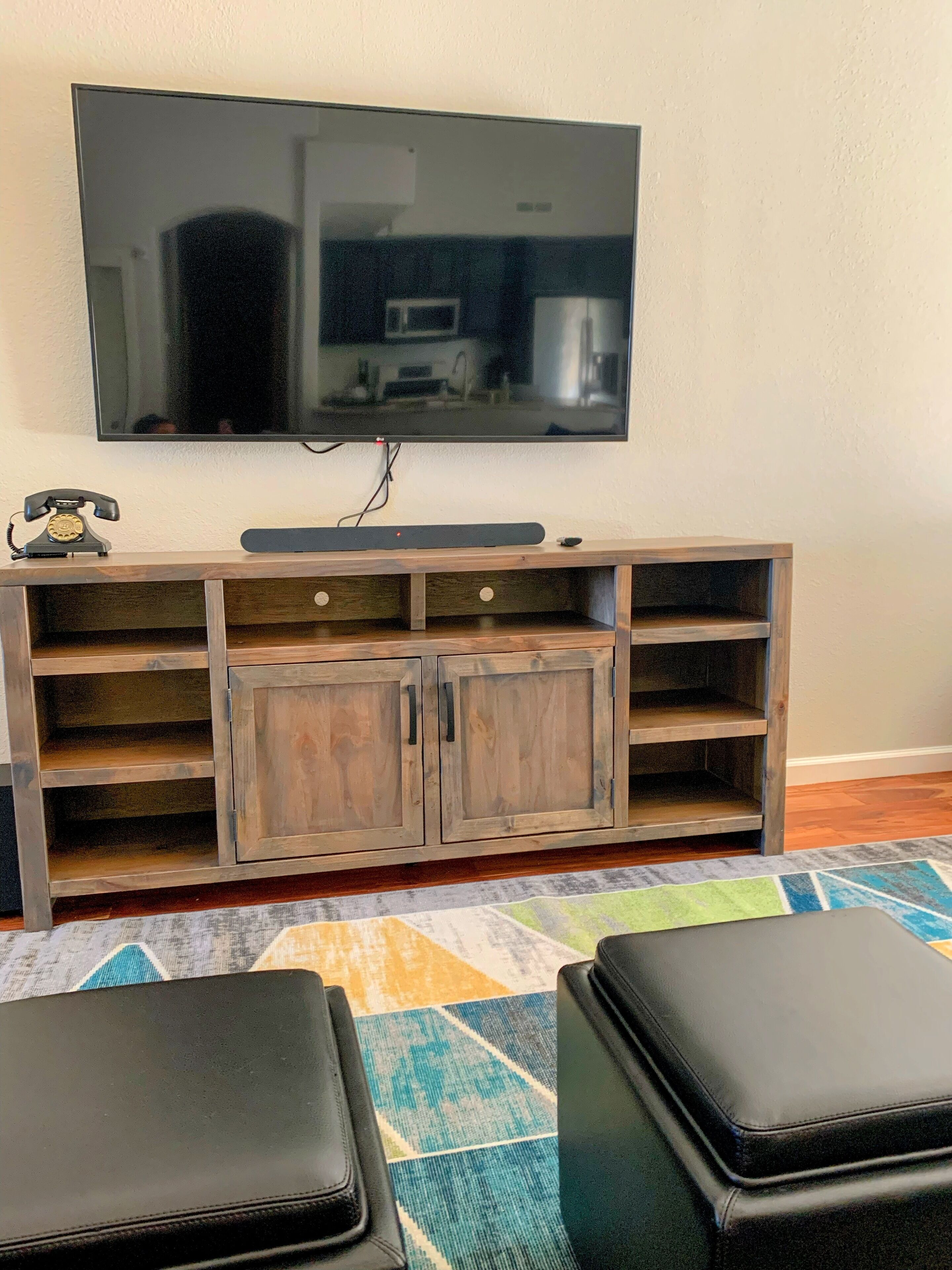 Uma sala de estar com televisão de tela plana fixada na parede, um móvel de madeira abaixo e dois pufes de couro preto sobre um tapete colorido.
