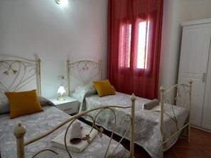 Ein Zimmer mit zwei Betten, jeweils mit weißem Metallgestell und einem gemusterten Bettbezug. Zwei Kissen, eines gelb und eines braun. Ein weißer Nachttisch mit Lampe steht zwischen den Betten. Ein roter Vorhang bedeckt teilweise ein Fenster.