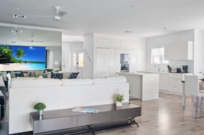 Uma sala de estar moderna com sofá, mesa de centro e televisão, integrada a uma cozinha com armários brancos e um espaço de jantar.