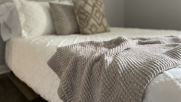 Una cama bien tendida con edredón blanco y una manta tejida gris.