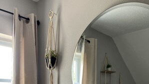 Un baño con un espejo redondo, un colgador de planta de macramé y una ventana con cortinas.