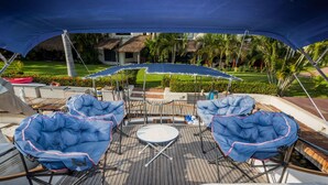 Un área de estar al aire libre con techo, con sillones reclinables y una mesa, rodeada por una terraza de madera y un edificio con techo de paja.