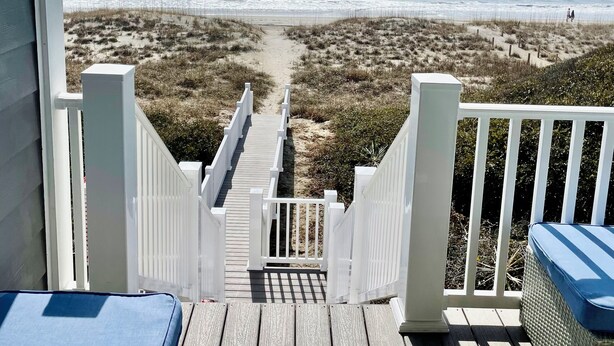 Una terraza de madera que da a una playa con un sendero arenoso y barandillas blancas.