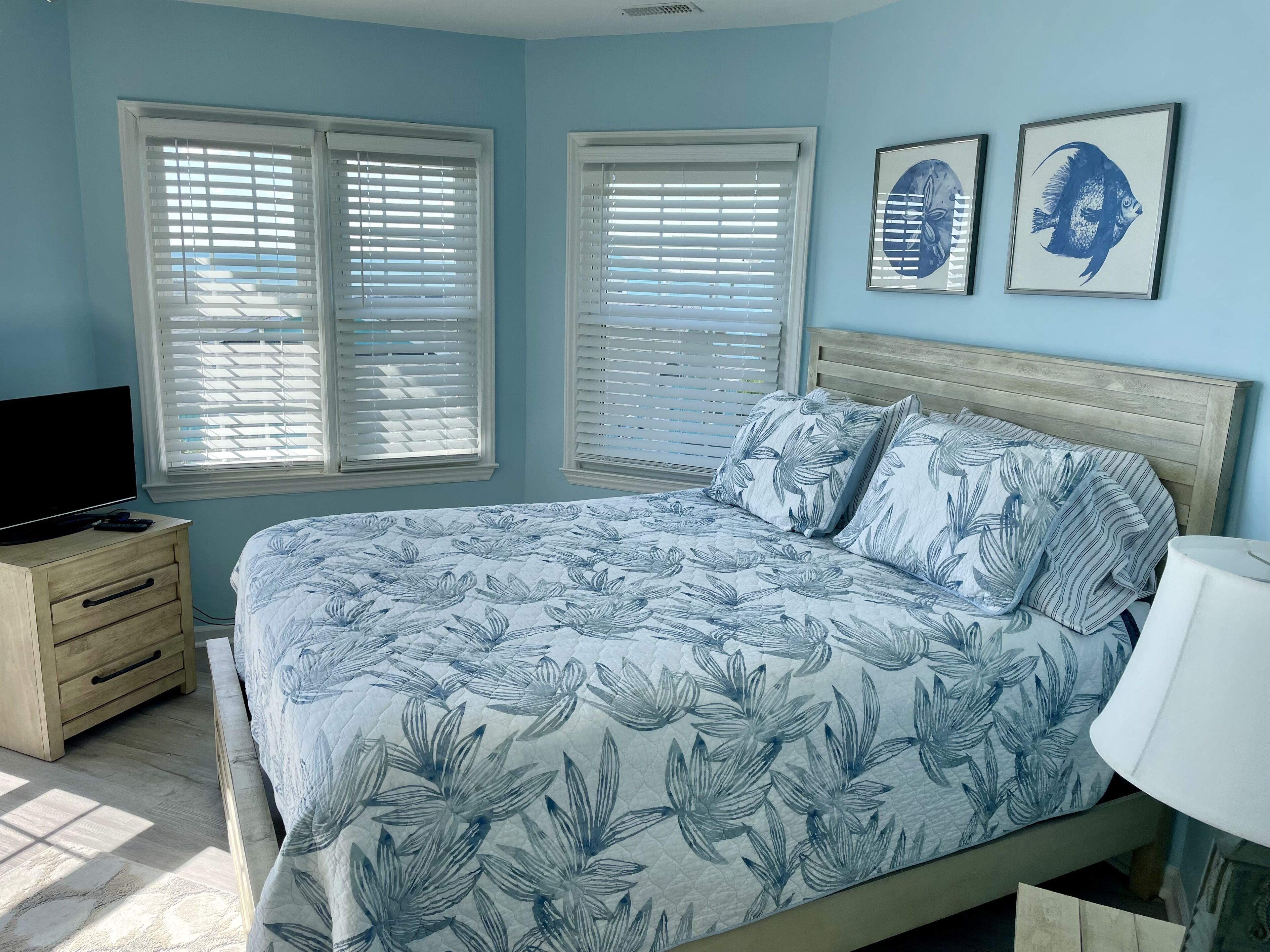 A bedroom with a bed, two windows with blinds, a TV on a wooden dresser, and two framed pictures on the wall.