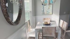 Un comedor con una mesa de madera rústica, sillas blancas, un espejo decorativo y una lámpara en el techo.