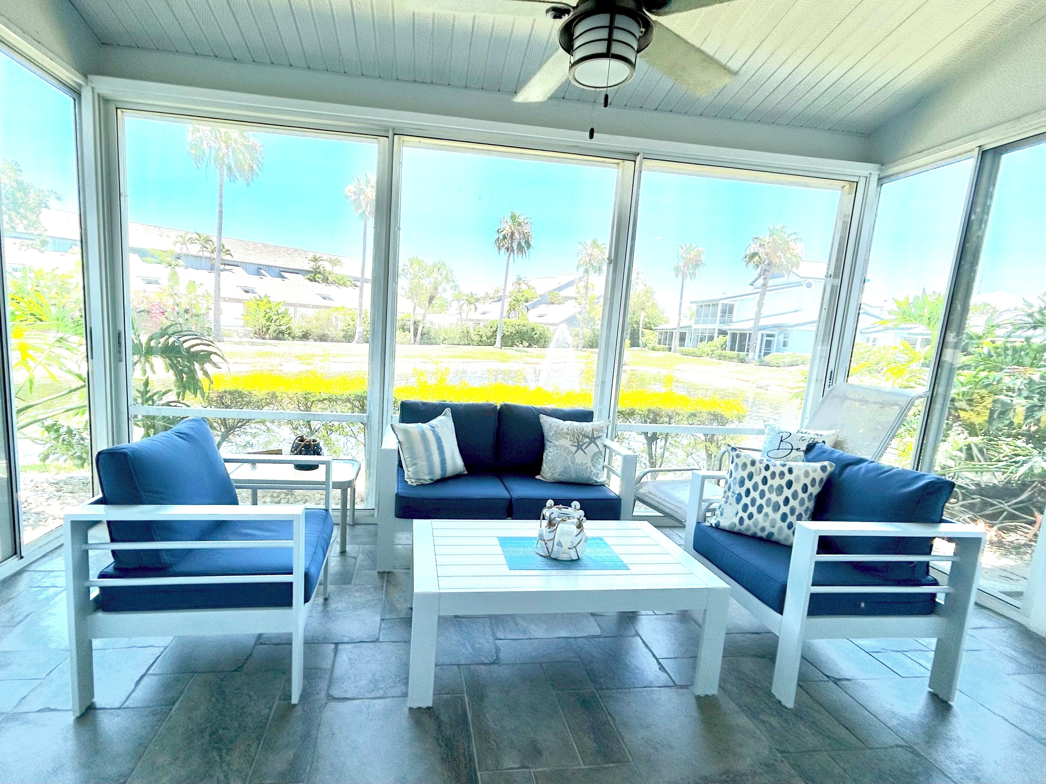 A sunroom with a white ceiling fan, large windows, a blue sofa, two white chairs, and a white coffee table.