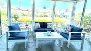 Une véranda avec un ventilateur de plafond blanc, de grandes fenêtres, un canapé bleu, deux chaises blanches et une table basse blanche.