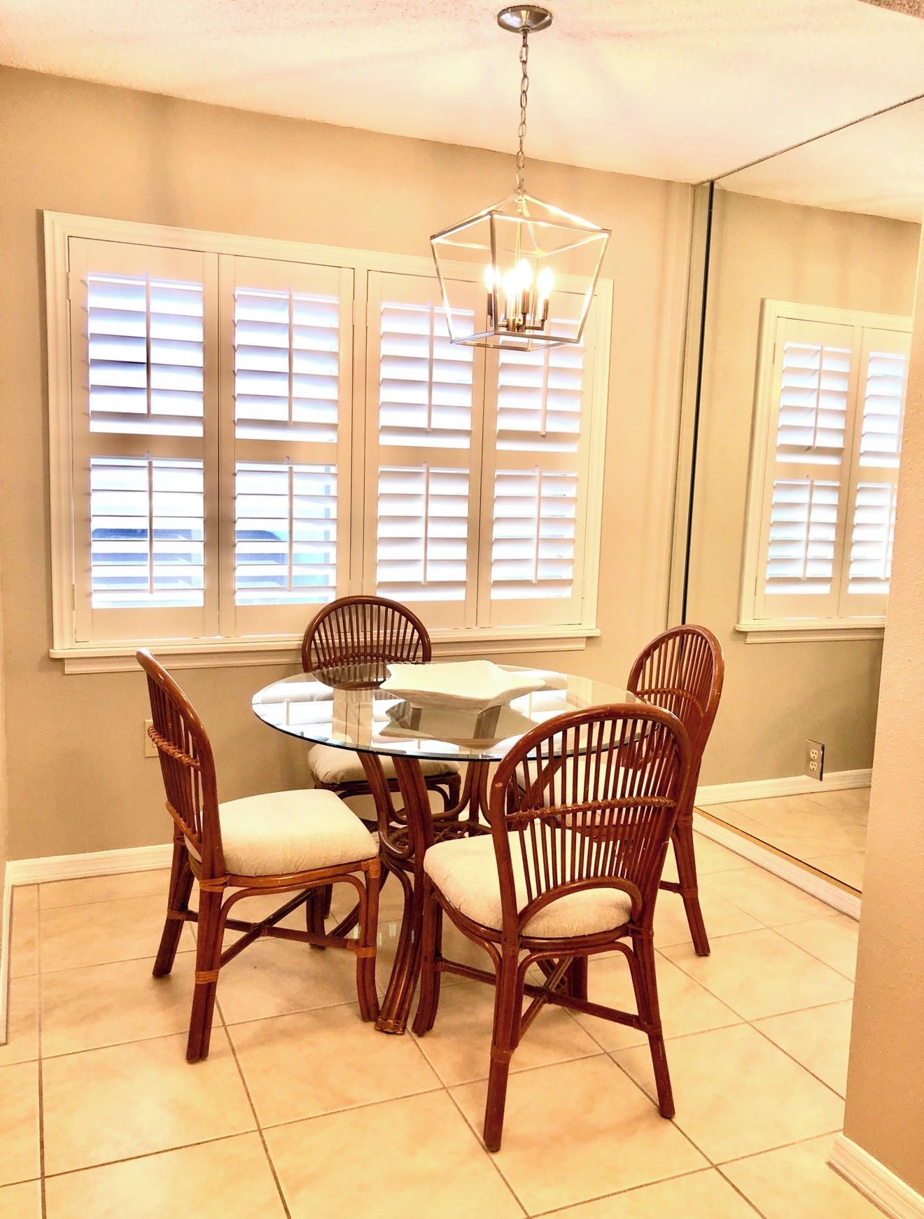 A dining area with a round glass table surrounded by wicker chairs, a hanging light fixture, and large windows with blinds.
