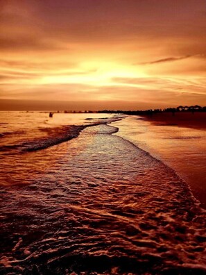 A beach with a long stretch of wet sand leading to the sea, under a sunset sky.