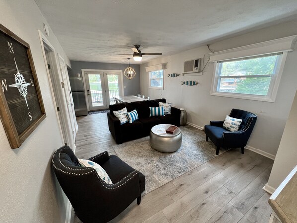 A spacious living room with a dark brown sofa, a blue armchair, and a round ottoman. There is a dining area in the background with a white table and chairs. - JOMO - Unit 2 TriTerrace *Relax Lakefront *Enjoy Fam (Rocky Mount)