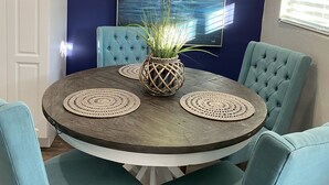 A dining area with a round table, four teal chairs, a woven centerpiece, and a large painting of a bird on the wall.
