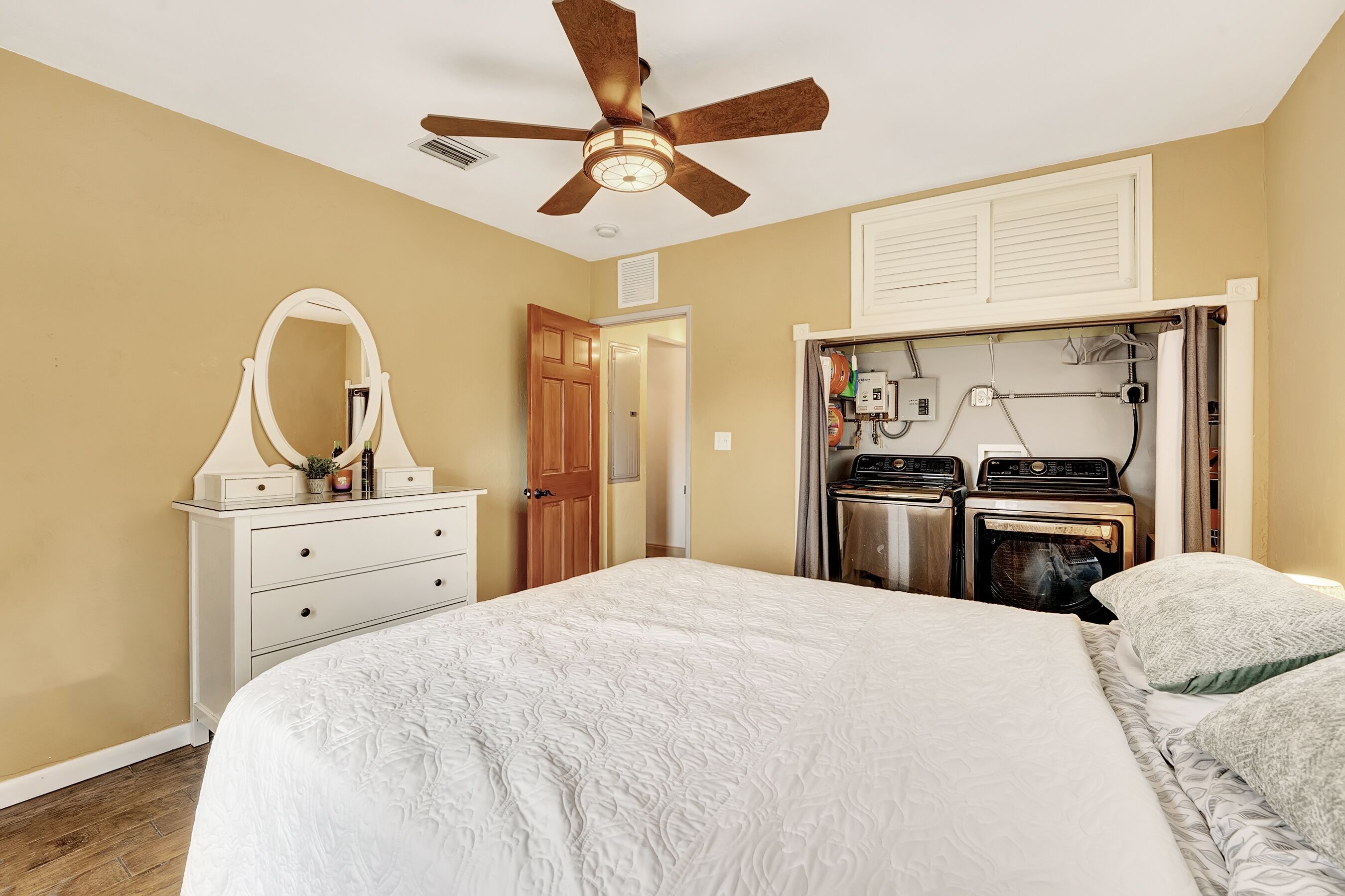 A bedroom with a bed, a ceiling fan, a dresser with a mirror, and a built-in washer and dryer.