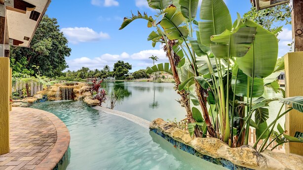 A tranquil indoor water feature with a brick pathway, lush tropical plants, and a clear blue sky. - Waterfront Oasis w/ Heated Pool and Spa, and Close to Beach! (Fort Lauderdale)