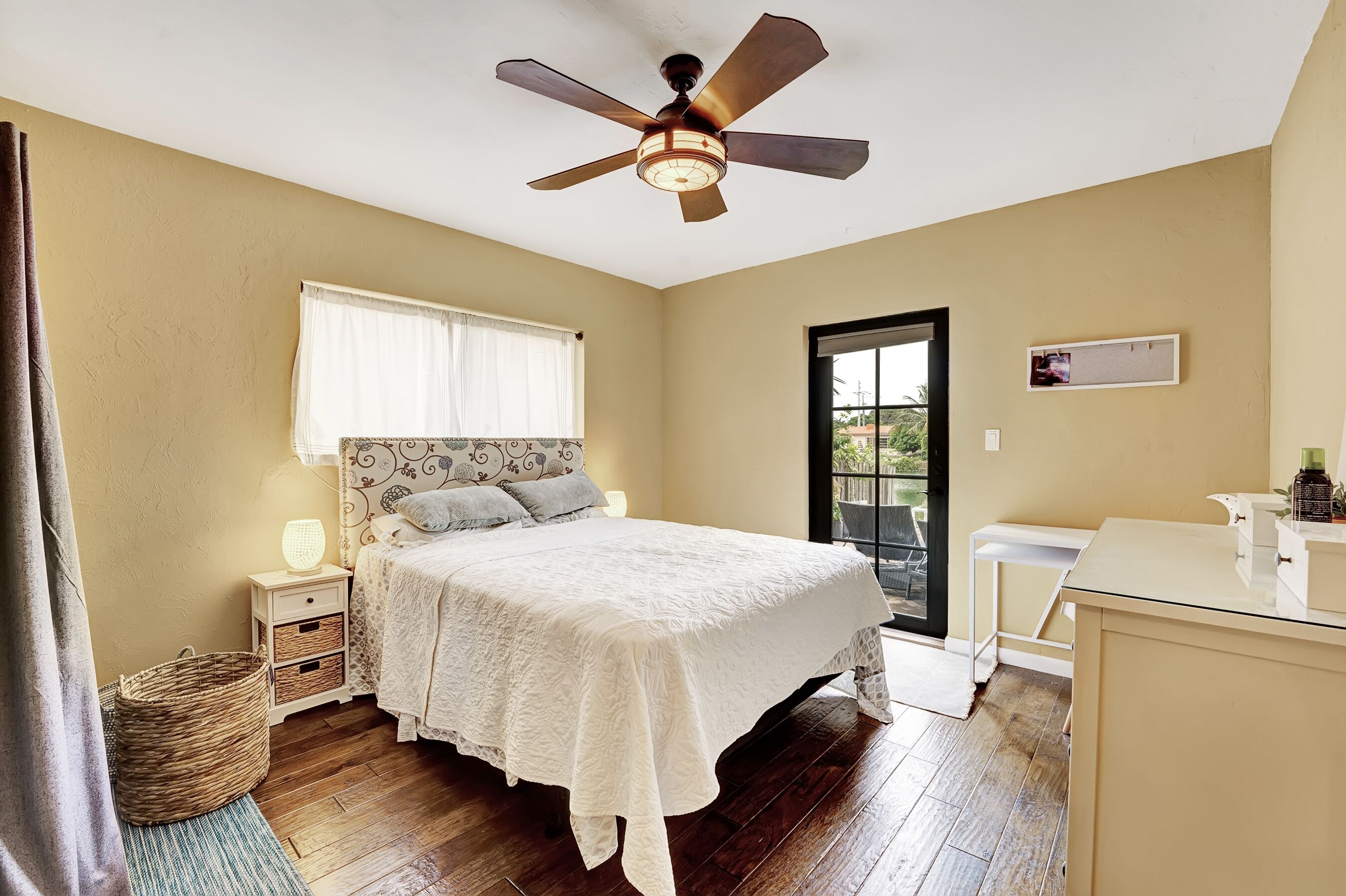 A bedroom with a bed, bedside tables, a ceiling fan, a window with curtains, a door, and a small table with a lamp.