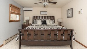 A bedroom with a wooden bed, a ceiling fan, an air conditioning unit, a window with blinds, and a wall-mounted picture.