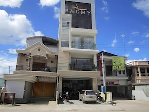 Un edificio de varios pisos con un llamativo letrero 'PALMY', un acceso cubierto y un visible letrero 'MADCO' en la planta baja.