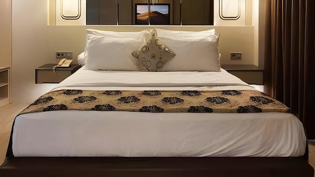 Cama bem arrumada com manta estampada, telefone de apoio e televisão fixada na parede.