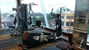 Ginásio com diversos equipamentos de exercício, um banco e vista para a cidade através de grandes janelas.