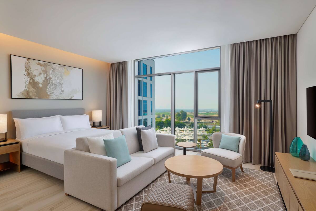 A modern hotel room with a large bed, a sofa, a small table, and a view of the city.